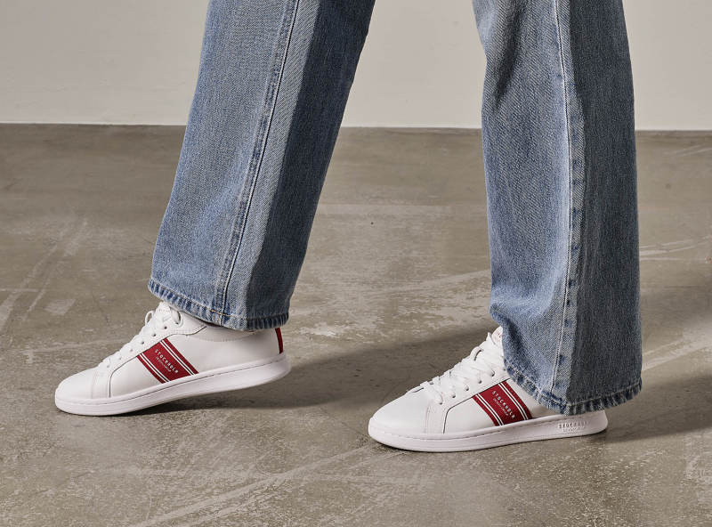 Hvite smale sneakers med rød detaljer på fot med jeans.